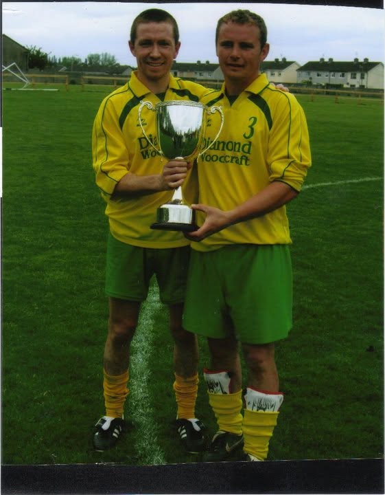 Two Crettyard United players in Diamond Woodcraft kit holding a trophy on the pitch